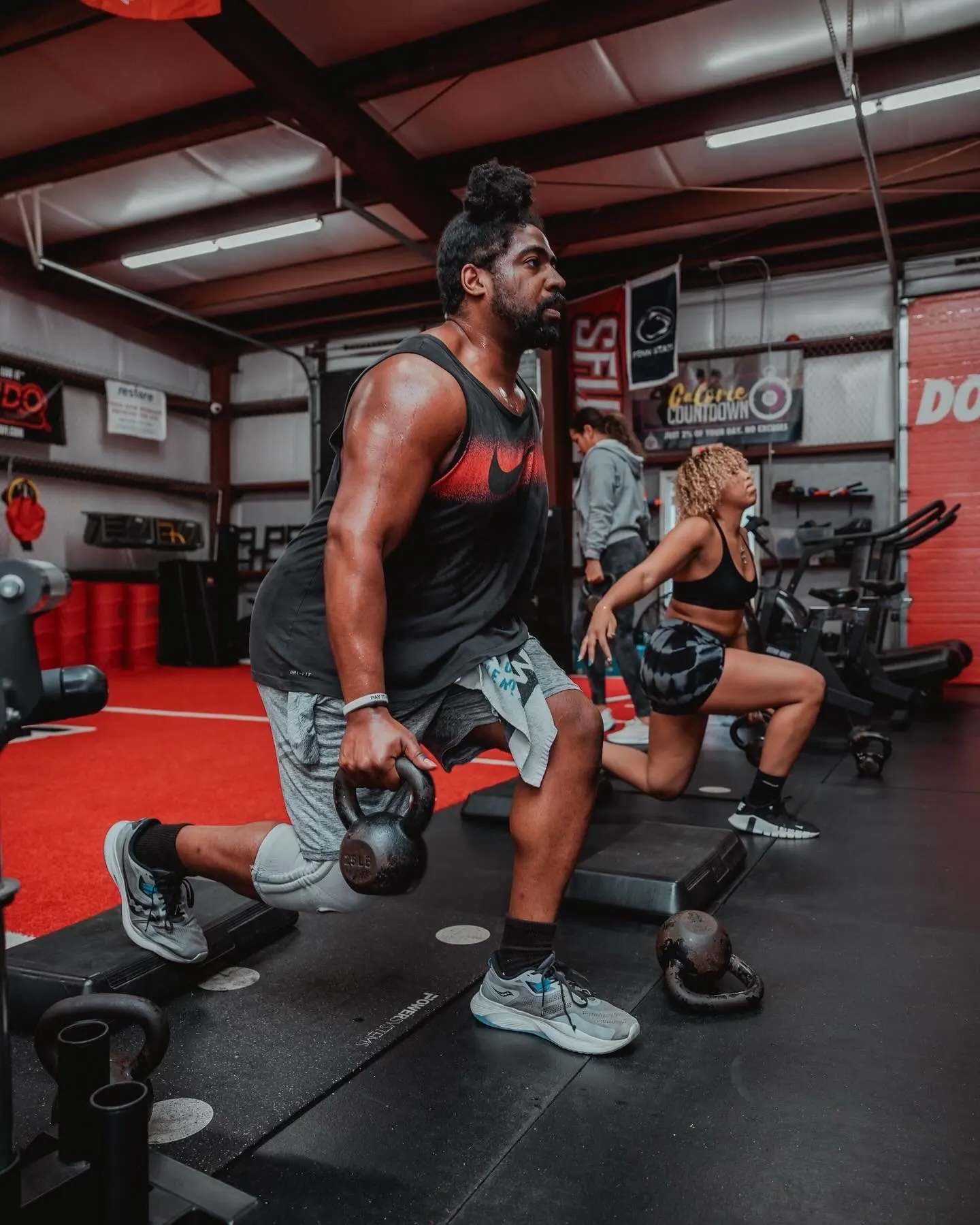 Adult members do kettlebell lunges side by side on the TNDO strength floor